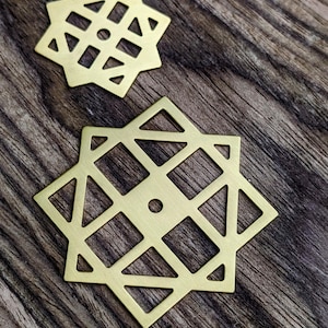 Peut inclure: Deux pièces décoratives en forme d'étoile de couleur laiton avec des découpes géométriques. Les pièces sont placées sur une surface en bois sombre et texturée. Les étoiles ont un carré central et des losanges environnants.