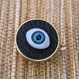 Puede incluir: Un objeto detallado en forma de ojo con iris azul y esclerótica blanca, colocado sobre una superficie negra texturizada dentro de un marco circular dorado. El iris del ojo presenta intrincados patrones azules.