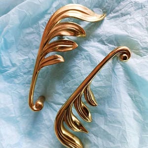 May include: Two gold-colored leaf-shaped cabinet pulls. Each pull features a detailed leaf design with multiple leaflets and a curved handle. The pulls are set against a light blue, textured background.