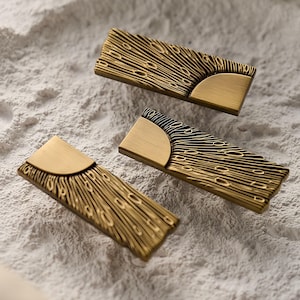 May include: Three brass-coloured cabinet pulls with a textured, linear design. Each rectangular pull features a semi-circular accent at one end. The pulls are arranged on a textured, off-white surface, showcasing their intricate details and warm colour.