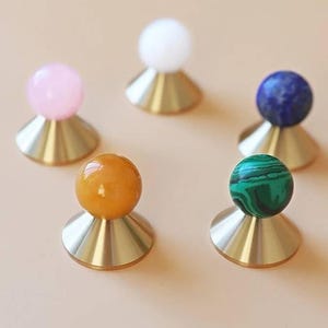 May include: Five decorative objects with brass bases and colourful spherical tops. The spheres are pink, white, orange, blue, and green. The objects are arranged on a light tan surface.