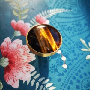 May include: A round, polished gemstone with a golden-brown, striped pattern, set in a gold-colored metal bezel. The gemstone is the focal point, resting on a teal surface with floral patterns.
