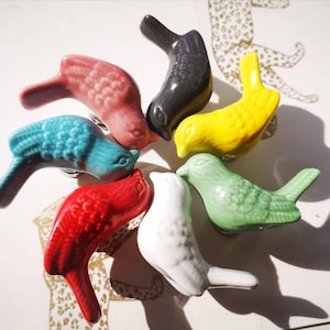 May include: A selection of ceramic bird-shaped cabinet knobs in a range of colours: red, teal, yellow, green, black, white, and pink. Each knob has a detailed bird design with a curved beak and tail, ideal for adding a decorative touch to furniture.