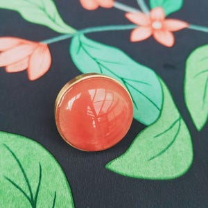 May include: A round, orange-red cabochon set in a gold-coloured metal bezel. The cabochon reflects light, and the background features a dark blue floral pattern with green leaves and coral-coloured flowers.