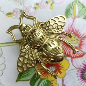 May include: A gold-colored bee-shaped brooch. The brooch has detailed wings and body segments, with a textured surface. The bee is set against a floral patterned background with pink, red, yellow, and green flowers.