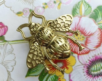 Puede incluir: Un broche dorado con forma de abeja. El broche tiene alas y segmentos corporales detallados, con una superficie texturizada. La abeja está sobre un fondo floral con flores rosas, rojas, amarillas y verdes.