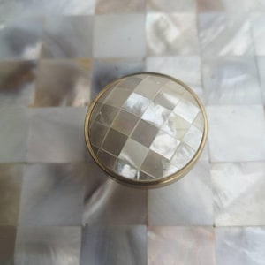 May include: A round, decorative cabinet knob with a faceted, mother-of-pearl center. The knob is set in a gold-toned metal frame. The background is a mosaic of square, iridescent tiles.