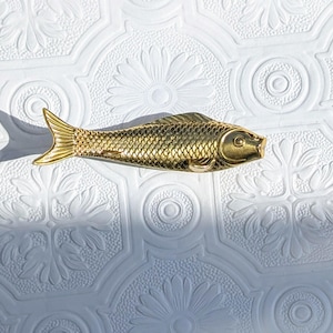 以下が含まれることがあります： 金色の魚の形をした装飾品。魚には詳細な鱗とひれがあり、花の模様の白い背景に配置されています。このオブジェクトは、おそらく家の装飾要素です。