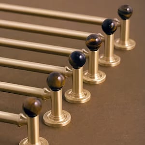 May include: Six gold-coloured towel rails with decorative finials. The finials are spherical, with a mix of brown and black tones. The rails are mounted on a matching gold-coloured base, creating a cohesive and elegant design.
