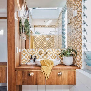 May include: A contemporary bathroom with a wooden vanity unit, a white basin, and a yellow towel. The walls are adorned with patterned tiles in yellow and white. A mirror reflects the room, and a hanging plant is visible.