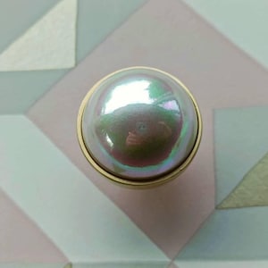 May include: A round, iridescent pearl-like knob with a gold-colored base. The knob reflects multiple colors and sits against a geometric background of pink, white, and green shapes. This decorative hardware is suitable for furniture.