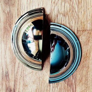 May include: Two semi-circular, silver-toned cabinet pulls with a decorative beaded edge, set against a light brown wood grain background. The pulls reflect the surrounding environment.