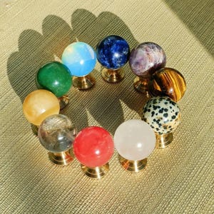 May include: A collection of ten gemstone spheres in various colors, including blue, green, yellow, red, and white, each mounted on a small gold-colored stand. The spheres are arranged in a circular pattern on a textured, golden-brown surface.