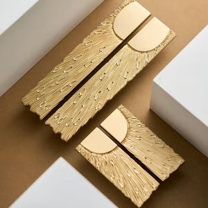 May include: Gold cabinet pulls with a textured, feather-like design. Each pull has a semi-circular accent and a rectangular base. The hardware is displayed on a neutral background, showcasing the intricate details and metallic finish.