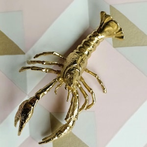 Könnte beinhalten: Eine goldfarbene Hummerfigur mit detaillierten Klauen, Beinen und Körpersegmenten. Der Hummer ist auf einer geometrischen Oberfläche mit rosa, weißen und goldenen Formen platziert. Die Figur hat eine strukturierte Oberfläche.
