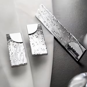 May include: Silver-toned metal accessories, including a rectangular bar and two smaller rectangular pieces, are arranged on a black and white surface. The pieces have a textured, patterned design and a curved, reflective top.