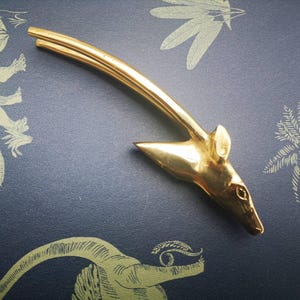 May include: A gold-colored metal bottle opener in the shape of a deer head. The opener has a curved handle and a pointed end. The deer head has detailed features, including ears and an eye. The opener is on a blue surface with a decorative pattern.
