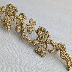 May include: A gold-colored, decorative cabinet pull in the shape of a stylized tree branch with detailed floral accents. The hardware is made of metal and has a textured finish. The pull is designed to be mounted horizontally.