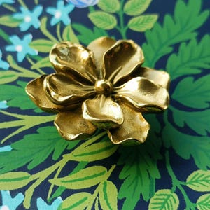 May include: A gold-coloured, flower-shaped decorative object. The flower has multiple petals and a textured surface. The background features a floral pattern with green leaves and blue flowers on a dark blue background.