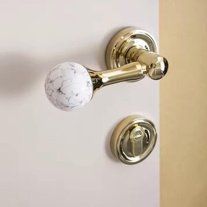 May include: A gold door handle with a white marble knob. The handle is attached to a white door.