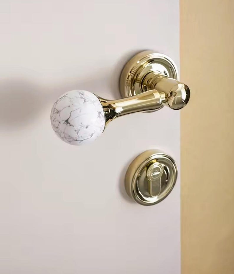 May include: A gold door handle with a white marble knob. The handle is attached to a white door.