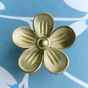 以下が含まれることがあります： 真鍮色の花の形をしたキャビネットノブ。5枚の花びらと中央の円形ディテールがあります。花びらにはエッチングされた線があります。青と白の花柄の背景に配置されています。