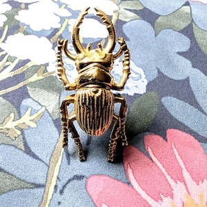 May include: A gold-colored beetle figurine with detailed legs, antennae, and body segments. The beetle is positioned on a floral patterned background with pink and blue flowers. The figurine is a decorative item.