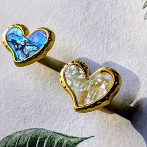 May include: Two heart-shaped earrings with iridescent abalone shells set in gold-toned metal. The earrings have a textured border and are displayed on a light background, showcasing their unique design and color variations.