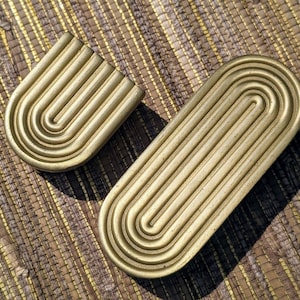 May include: Two gold-colored, decorative trays with a ridged, curved design. One tray is U-shaped, and the other is an elongated oval. The trays are placed on a woven, textured surface.