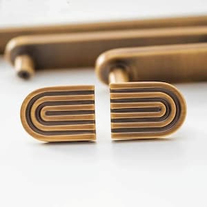 Puede incluir: Primer plano de herrajes de armario de bronce antiguo. Los herrajes presentan una forma rectangular redondeada con una serie de líneas curvas. Los herrajes son de color bronce cálido y parecen ser de metal. Los herrajes están diseñados para armarios y cajones.