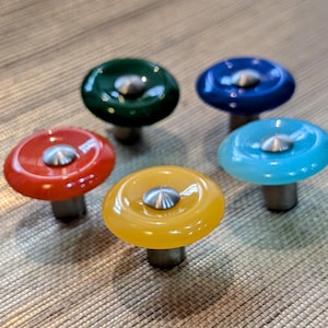 May include: Five cabinet knobs in various colours: red, green, blue, yellow, and turquoise. Each knob features a rounded, glossy top with a silver centre and a metal base. These decorative knobs are likely for furniture.