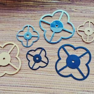 May include: Six flower-shaped decorative cutouts in varying sizes and colours. The cutouts are in shades of blue and beige, with a central circular element and four curved petals. They are arranged on a textured wooden surface.