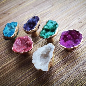 May include: Six geode cabinet knobs in various colours. Each knob has a unique crystal formation in shades of blue, purple, green, red, and white, with gold-coloured edges and wooden bases. These decorative knobs add a touch of natural beauty.