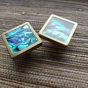 Puede incluir: Dos tiradores cuadrados decorativos para armarios con incrustaciones de concha de abulón iridiscente. Los tiradores tienen un marco de metal dorado y están colocados sobre un fondo marrón texturizado. La concha de abulón muestra tonos de azul, verde y negro.