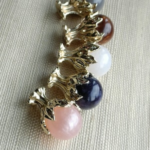 Puede incluir: Cinco cuentas de cabujón decorativas en fila. Cada cuenta tiene una base dorada ornamentada. Las cuentas son en tonos de gris, marrón, blanco, azul oscuro y rosa. El fondo es una tela beige claro.