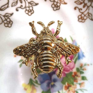 May include: A gold-colored bee-shaped brooch with detailed wings and body segments. The brooch is set against a floral patterned background with pink, blue, and yellow flowers.