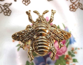 Könnte beinhalten: Goldfarbene, bienenförmige Brosche mit detaillierten Flügeln und Körpersegmenten. Die Brosche befindet sich vor einem floralen Hintergrund mit rosa, blauen und gelben Blumen.