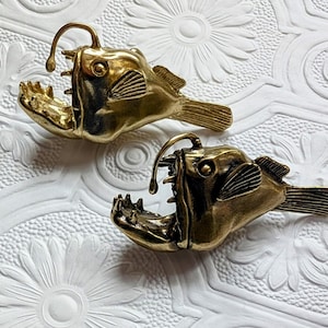 May include: Two brass anglerfish sculptures with open mouths and detailed fins are displayed on a white, textured surface. The sculptures have a vintage aesthetic, with a golden hue and intricate detailing.