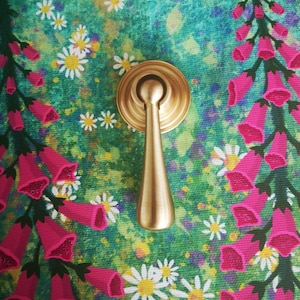 以下が含まれることがあります： 涙の形をしたハンドルが付いた金色のキャビネットプルが、花の背景に配置されています。背景には、ピンクのジギタリス、白いデイジー、緑とターコイズブルーの塗装キャンバスがあります。