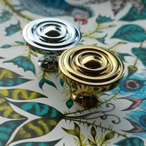 May include: Two decorative cabinet knobs, one silver and one gold, with a circular design. The knobs are set against a colourful floral and peacock feather patterned background.