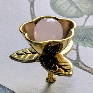 May include: A decorative, gold-coloured floral-shaped cabinet knob. The knob features a light pink, oval-shaped stone set within a flower-like cup. The base has a leaf design and a screw for attachment. The background has a floral and bird pattern.