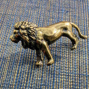 May include: A detailed brass lion figurine, showcasing a regal pose with its mane and muscular form. The sculpture is a warm gold color, set against a blue and tan woven background. This decorative piece could be used as a paperweight or a shelf decoration.