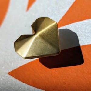 May include: A heart-shaped, brass-colored object with a faceted design. The object is casting a shadow on a surface with orange and white geometric patterns. The object has a brushed metal finish.
