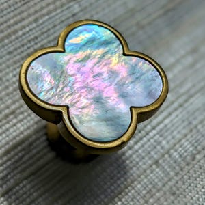 May include: A decorative cabinet knob with a four-leaf clover design. The knob features a shimmering, iridescent mother-of-pearl center framed by a gold-colored metal. The clover shape is a classic design element.