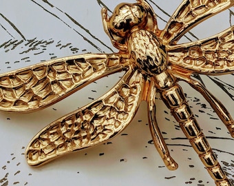 Puede incluir: Broche de libélula dorada con alas y cuerpo detallados. El broche tiene una superficie texturizada y está sobre un fondo blanco con un sutil patrón de hojas. El diseño de la libélula es muy detallado.