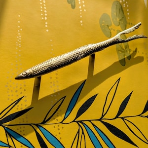 Könnte beinhalten: Ein dekorativer, goldfarbener Fischgriff. Der Griff ist auf zwei kleinen Pfosten montiert und wirft einen Schatten auf einen gelben Hintergrund mit einem Blatt- und Blumendesign. Der Fisch hat ein strukturiertes Schuppenmuster.