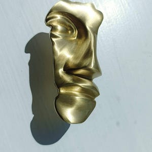 May include: A gold-coloured sculpture of a face, with a smooth, flowing design. The sculpture is highly detailed, with visible features like a nose, lips, and an eye. The piece casts a shadow on the white surface below.