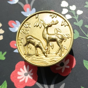 Könnte beinhalten: Ein goldfarbener, kreisförmiger Knopf mit einem detaillierten Relief von zwei Hirschen in einer natürlichen Umgebung. Ein Hirsch steht, der andere grast. Der Knopf hat einen erhabenen Rand und ist vor einem floral gemusterten Hintergrund platziert.