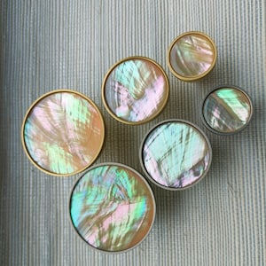 May include: Six circular cabinet knobs with iridescent mother-of-pearl inlays. The knobs have gold and silver-toned metal rims. The knobs are displayed on a light blue and white striped background.