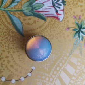 Könnte beinhalten: Ein runder, irisierender Schrankknopf mit einem goldfarbenen Metallrand. Die Oberfläche des Knaufs wechselt von Blau zu Rosa und Gelb. Er liegt auf einem gelben Hintergrund mit floralen Mustern.